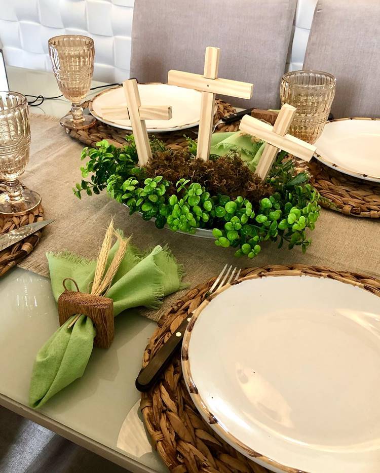 Uma mesa de jantar decorada para a Páscoa Cristã: nela contém pratos, talheres, sousplat, porta-guardanapos, taças e decoração com cruzes de madeira. As cores são marrom, branco e verde.