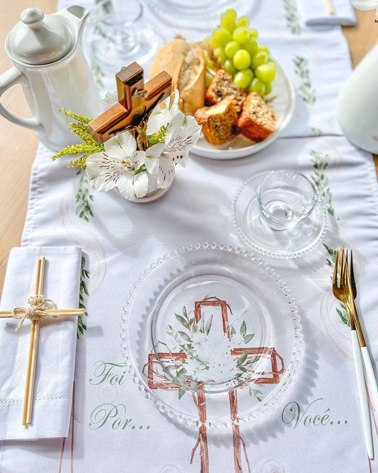 Uma mesa de jantar decorada para a Páscoa Cristã: toalha branca com a frase "Foi por você", prato e xícara de vidro, talheres dourados, bule de cerâmica, guardanapo com cruz de palito amarrado, bandeja com pães, bolos e uvas verdes, decoração com flores e cruz com imagem de Jesus Cristo.