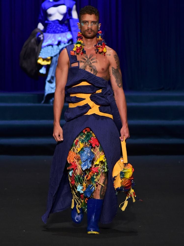 Homem de pele morena desfilando com vestido azul com detalhe em amarelo e outros detalhes coloridos