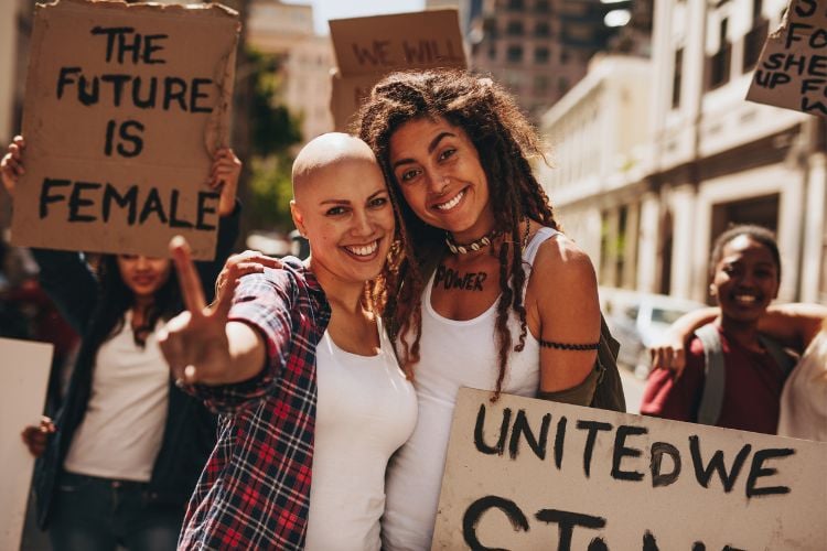 Duas mulheres abraçadas: uma careca e uma negra de dreads. elas usam regata branca e estão em um protesto feminino com outras mulheres segurando placas com frases feministas
