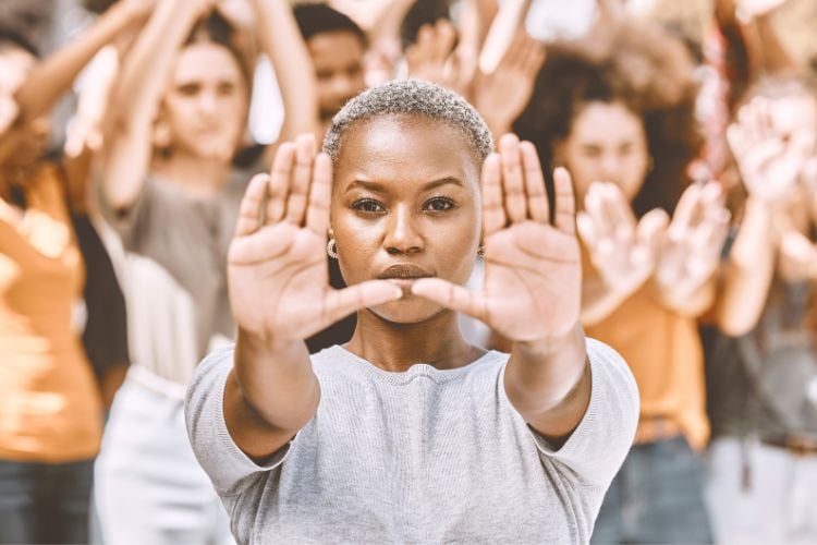 Mulher negra em protesto  fazendo sinal com as mãos ao fundo outras mulheres fazendo o mesmo sinal 