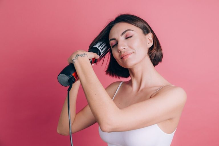 Foto de fundo rosa com mulher de pele clara usando regata branca e secando o cabelo curto e castanho com escova secadora