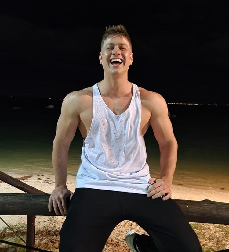 Ex-bbb Lucas Gallina de regata branca sentado em cerca de madeira de frente à praia à noite
