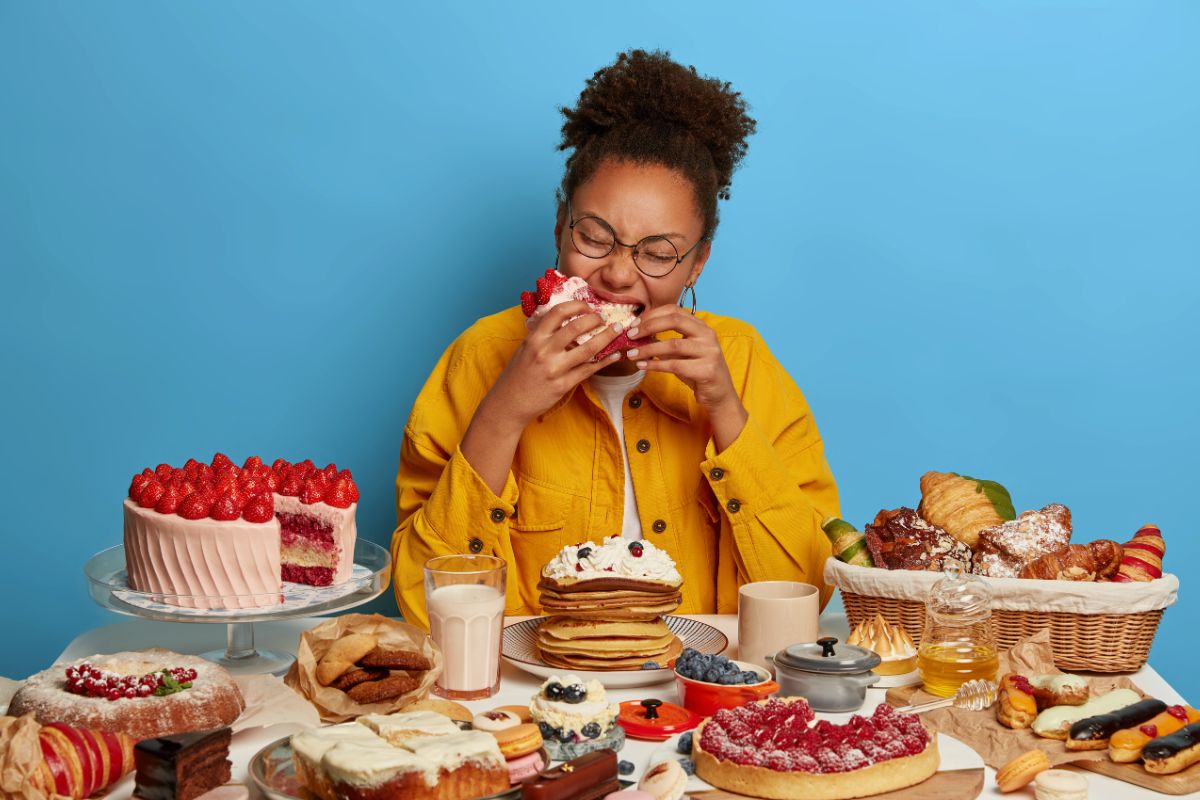 Imagem de uma mulher de pele negra usando blusa amarela comendo doce em uma mesa repleta de doces