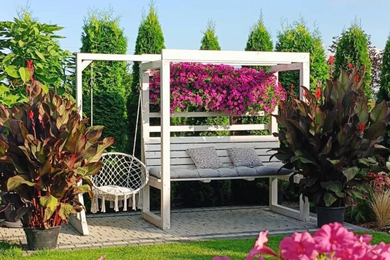 A imagem retrata um ambiente de jardim com estrutura em madeira branca, criando um espaço de descanso. Há um balanço de macramê ao lado e um banco acolchoado com almofadas. Plantas vibrantes e flores roxas embelezam o local.