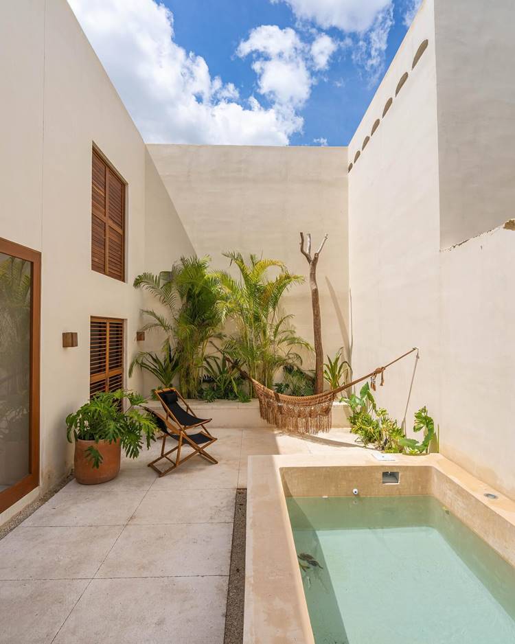 Área externa de casa com piscina e pequeno jardim na parede de fundo, com plantas tropicais. Há uma rede, cadeiras e outras plantas em vasos