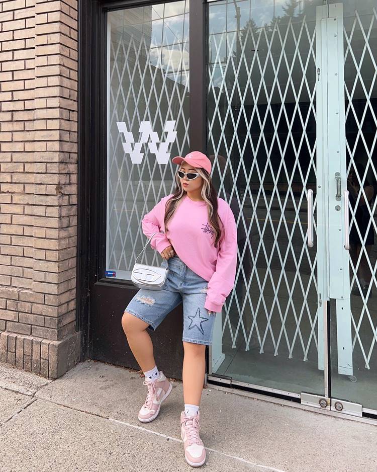 Garota ocidental usando jorts com bordado, tênis da Nike, moletom rosa e boné rosa, além de bolsa prateada, em frente à porta grande de um local público.

