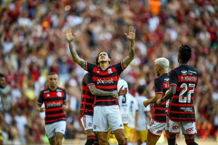 Foto do atacante comemorando gol com os braços para cima cercado de seus companheiros de time