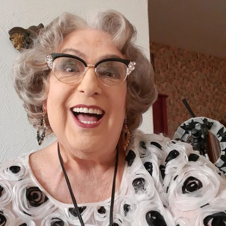 Selfie de Mamma Bruschetta com óculos pretos, maquiada e usando uma roupa com bordado de flor em casa.
