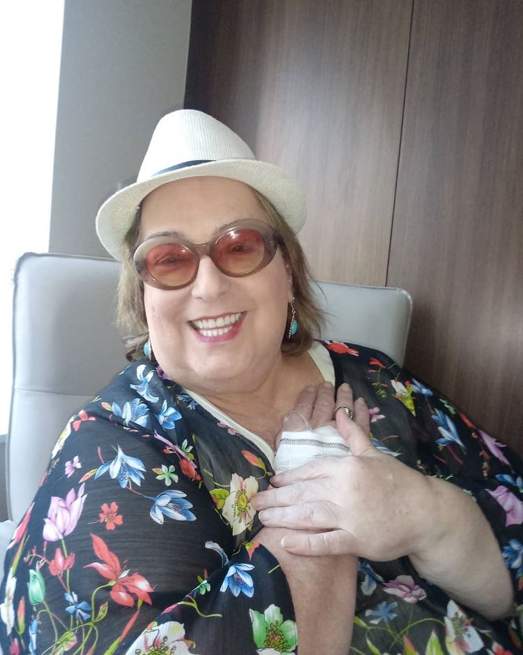 Mamma Bruschetta com chapéu, vestido floral, sentada em uma cadeira com as mãos no peito, uma delas com curativo.