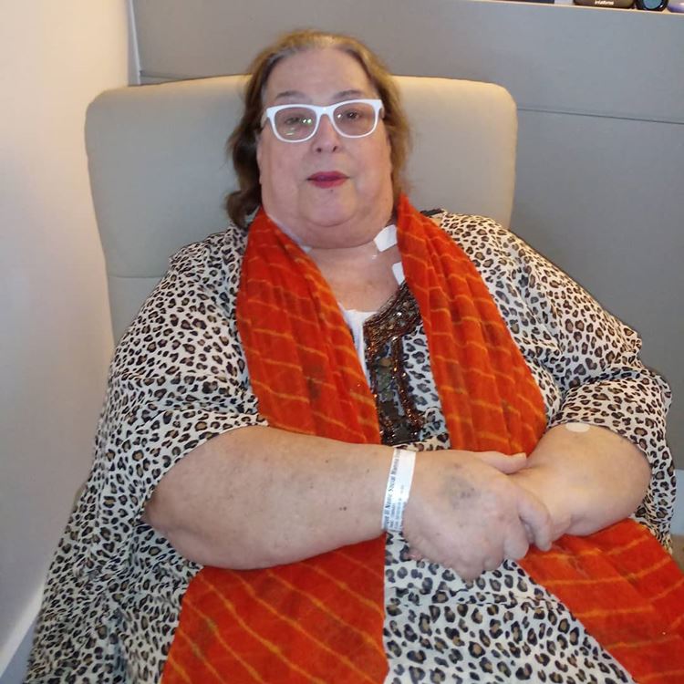 Foto da apresentadora usando um vestido com estampa de onça, cachecol laranja e óculos brancos, sentada em uma poltrona.
