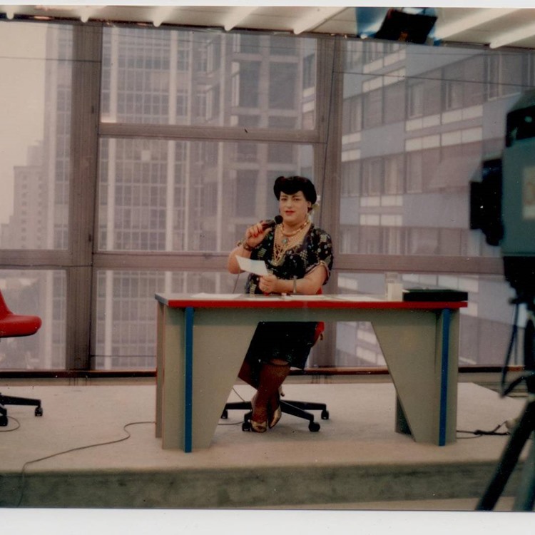 A personagem apresentando um programa antigo, usando um vestido preto e sentada em uma cadeira em frente a uma mesa.
