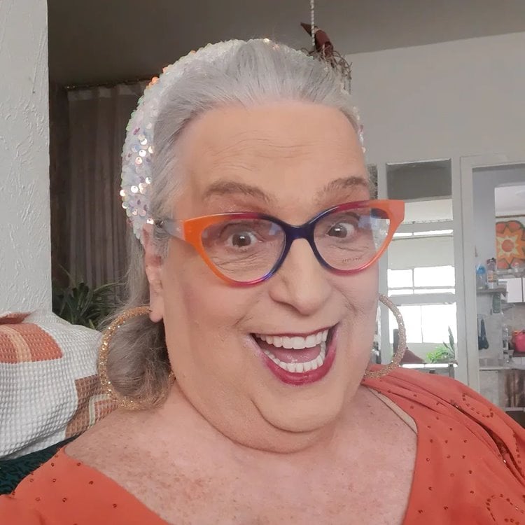 Selfie de Mamma Bruschetta com óculos coloridos e roupa laranja, sentada em um sofá em casa.