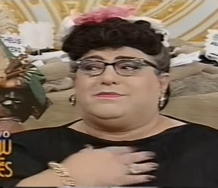 A personagem em um programa antigo, usando óculos e roupa preta.
