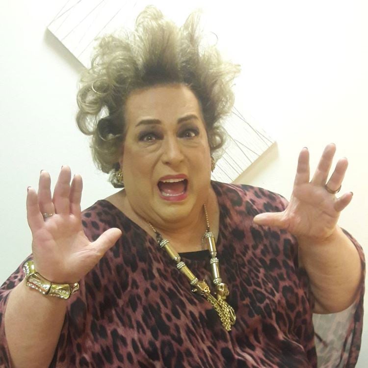 Foto de Mamma Bruschetta com semblante assustado, mãos erguidas, usando uma roupa com estampa de onça, joias douradas e cabelo arrepiado.
