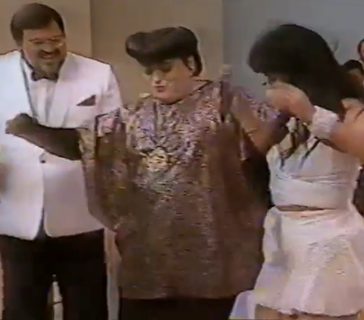 A personagem em um programa antigo, usando uma roupa brilhante dourada ao lado de Gretchen e outra para o computador.