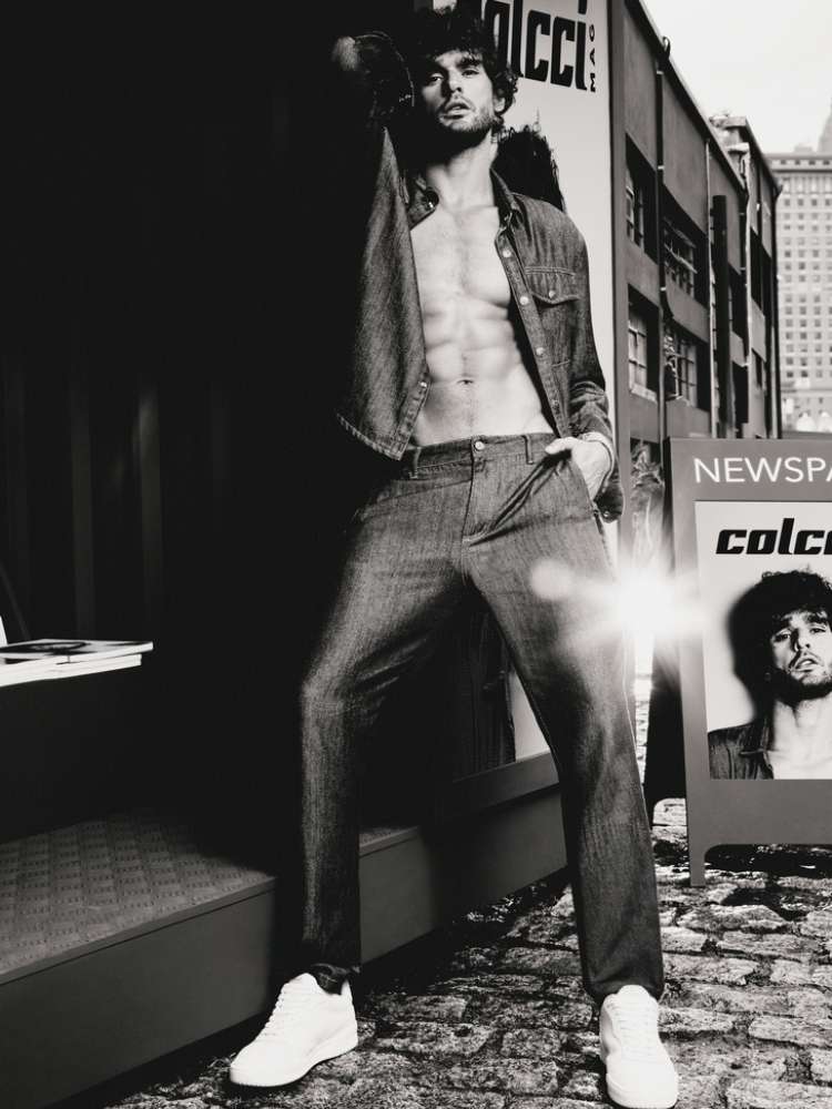 Marlon Teixeira de jaqueta e calça jeans ao lado de um cartaz com seu rosto e a logo da marca "Colcci". A foto é em preto e branco