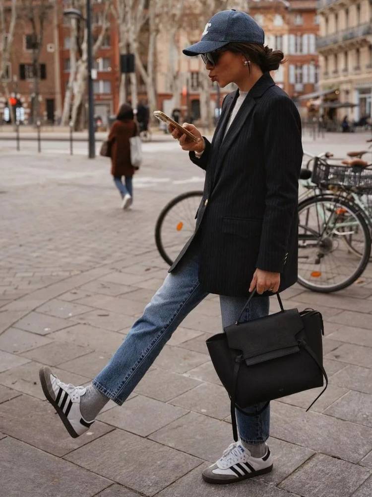 Mulher de lado andando na rua usando boné cinza, óculos de sol, calça jeans curta, blazer preto e sapatênis branco, preto e bege