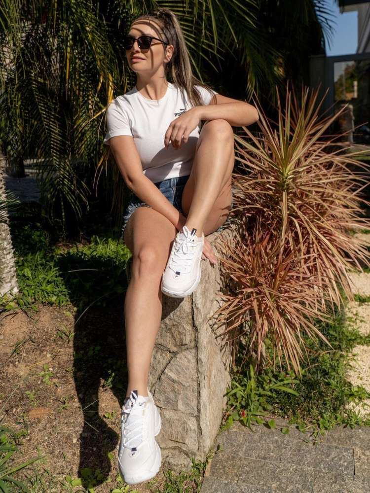 Mulher sentada em muro de pedras usando shorts jeans, blusa branca e tênis esportivo branco