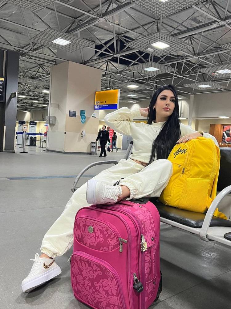 Mulher de pele clara e cabelo preto sentada no aeroporto, com mochila amarela e mala rosa, usando conjunto claro de moletom e tênis branco plataforma