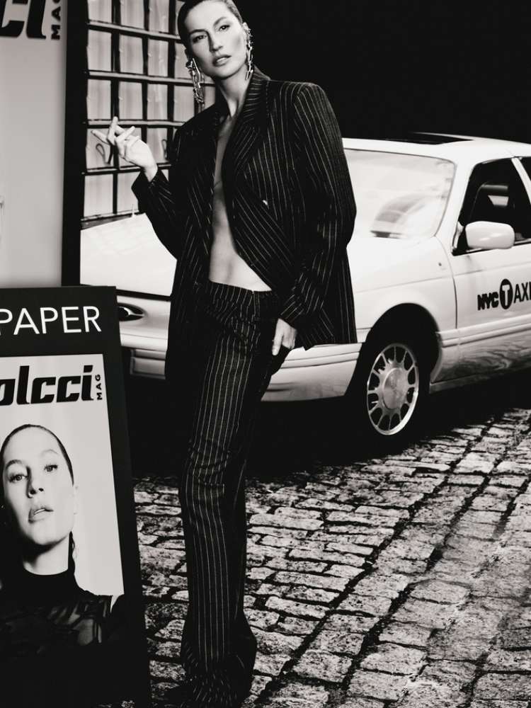 Foto em preto e branco de Gisele Bündchen usando blazer e calça em frente a um taxi e ao lado de um cartaz com seu rosto e o escrito "colcci"