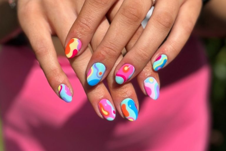 A imagem mostra unhas decoradas com nail art colorida e psicodélica, combinando tons vibrantes como azul, rosa, amarelo e laranja. Os desenhos têm formas abstratas e fluidas, criando um visual artístico, moderno e alegre.