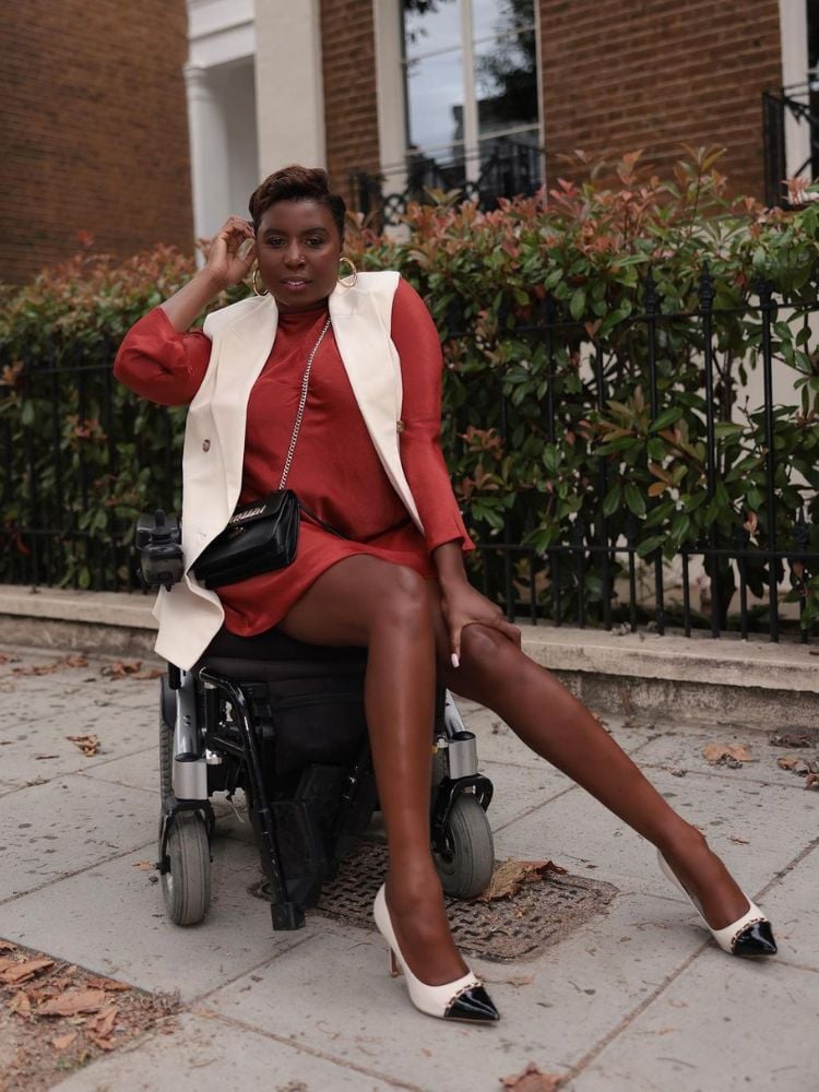 mulher em cadeira de rodas usando vestido marrom, colete de alfaiataria branco e scarpin em duas cores