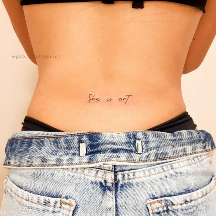 foto das costas de uma mulher com a tatuagem da frase She is art, Ela é arte em inglês