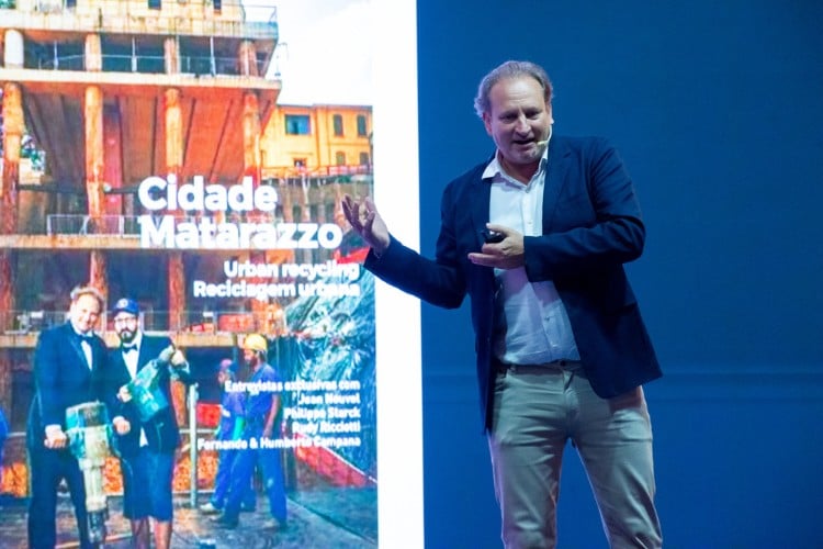 Alex Allard de terno azul, camisa branca e calça bege sobre palco de evento e slide da Cidade Matarazzo ao fundo