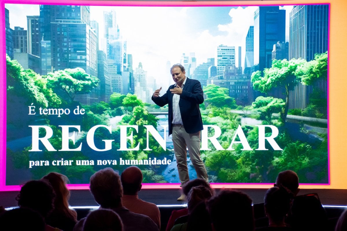 Alex Allard usando terno azul, camisa clara e calça bege dando palestra em palco com a foto de cidade com árvores ao fundo e a palavra "regenerar" junto no telão