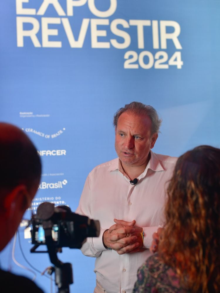 Alex Allard de camisa clara falando próximoa microfones e parede azul da Expo Revestir 2024 ao fundo