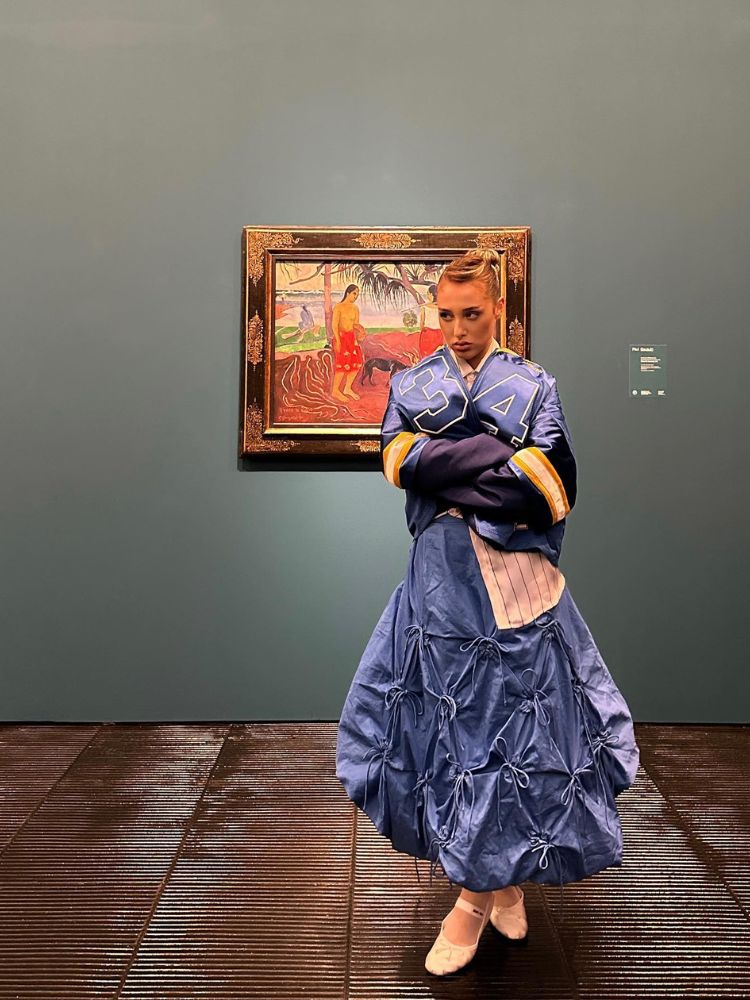 Malu Borges em museu usando saia longa azul balonê com laços, jaqueta azul e sapatilha