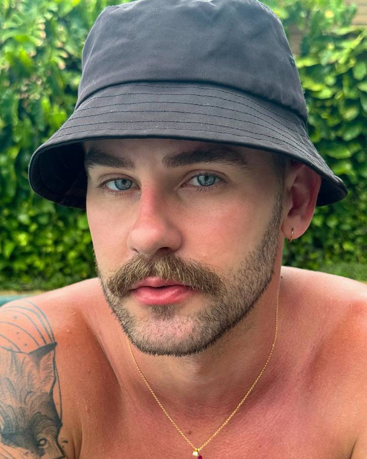 Homem com barba estilo beardstache, uma das tendências de 2024. Ele está sem camisa, com joias douradas e usa chapéu de pescador preto