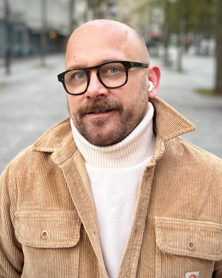 Homem com barba estilo beardstache, uma das tendências de 2024. Ele está usando roupa de inverno nas cores off white e bege, de óculos, fones de ouvido em rua pública