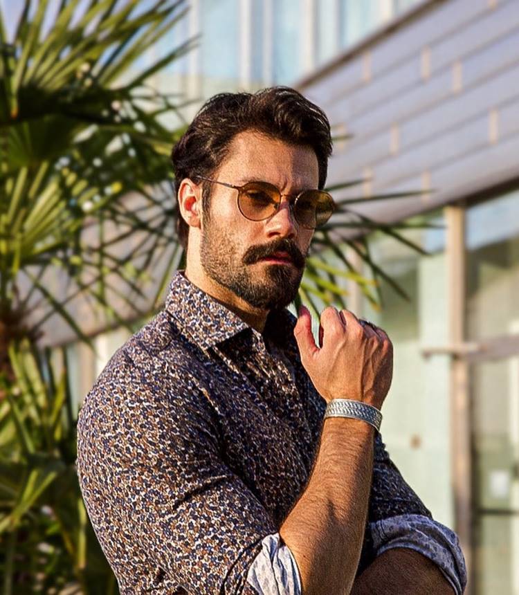 Homem com barba estilo beardstache, uma das tendências de 2024. Ele está usando camisa floral, com joias, óculos de sol, em local público