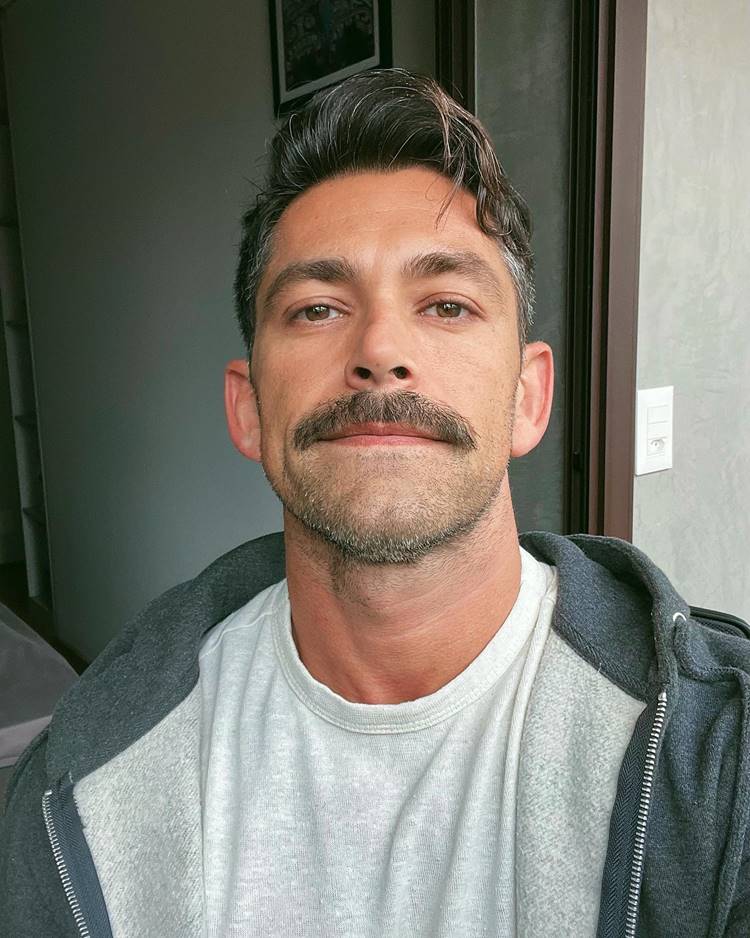 Homem com barba estilo beardstache, uma das tendências de 2024. Ele está com camisa e blusa de frio dentro de casa