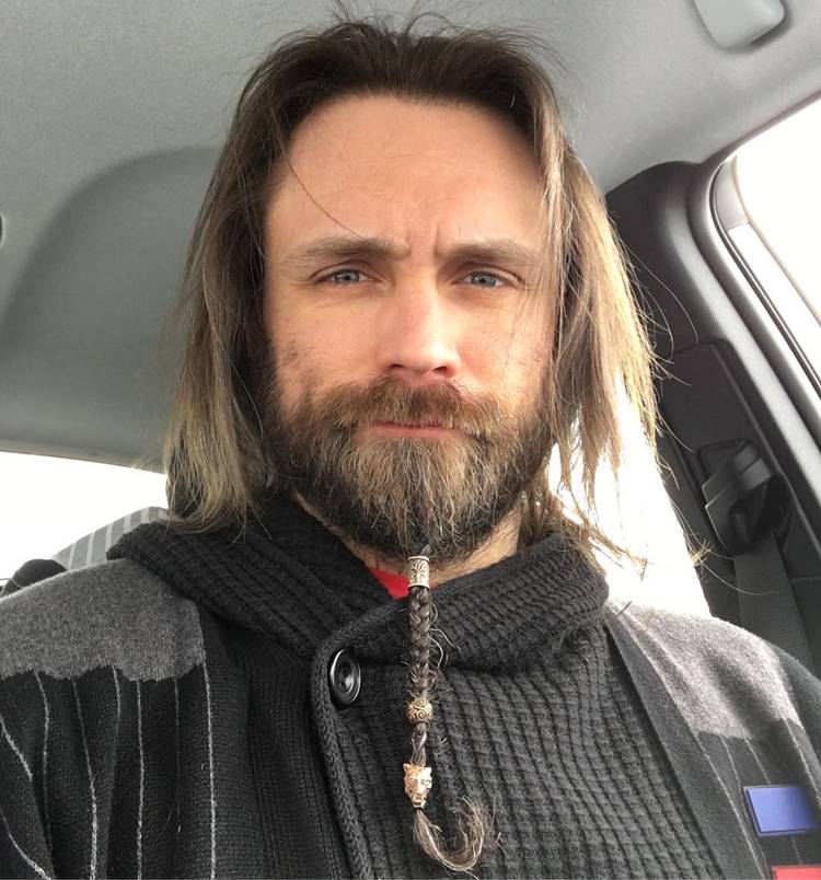 Homem com pelo do rosto trançada. Ele tem o cabelo longo, está com roupa de frio dentro de carro