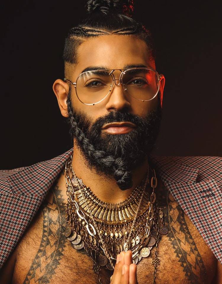 Homem com pelo do rosto trançada. Ele tem o cabelo longo que está trançado e amarrado, está com casaco xadrez, usa colares, óculos e está em fundo preto