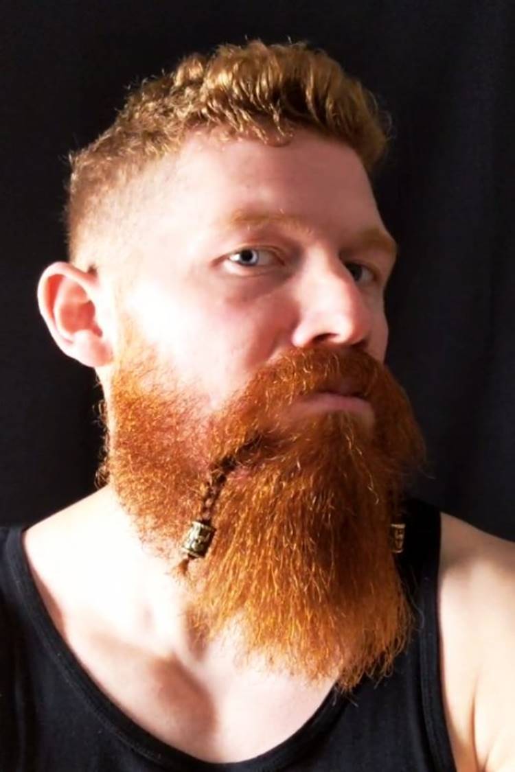 Homem com pelo do rosto trançada. Ele tem o cabelo ruivo, está usando regata preta em fundo preto