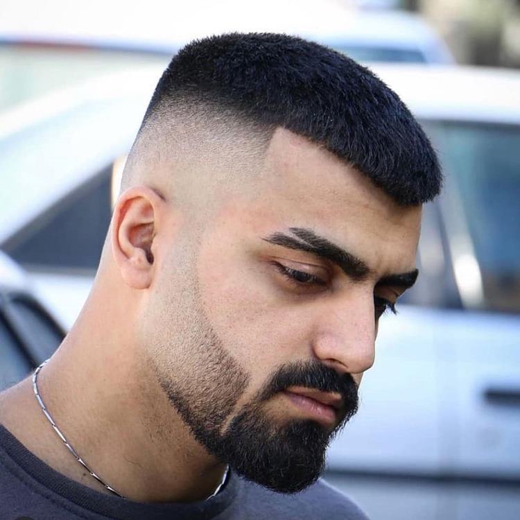 Homem com barba degradê, uma das tendências de 2024. Ele usa roupa preta, corrente no pescoço e está próximo a um carro