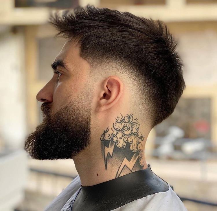 Homem com barba degradê, uma das tendências de 2024. Ele usa roupão de barbearia, tem tatuagem no pescoço e está na barbearia 