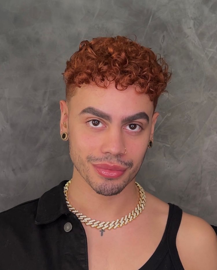 Homem com barba por fazer marcada, uma das tendências de 2024. Ele usa roupa preta, joias douradas e está em fundo cinza