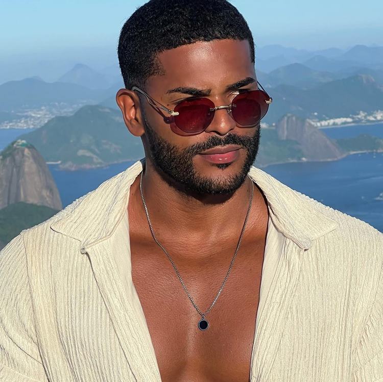 Homem com barba por fazer marcada, uma das tendências de 2024. Ele usa roupa branca, corrente e está no Rio de Janeiro