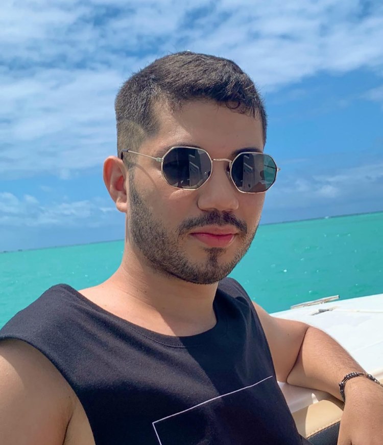 Homem com barba por fazer marcada, uma das tendências de 2024. Ele usa roupa preta, óculos de sol e está em uma embarcação em alto mar