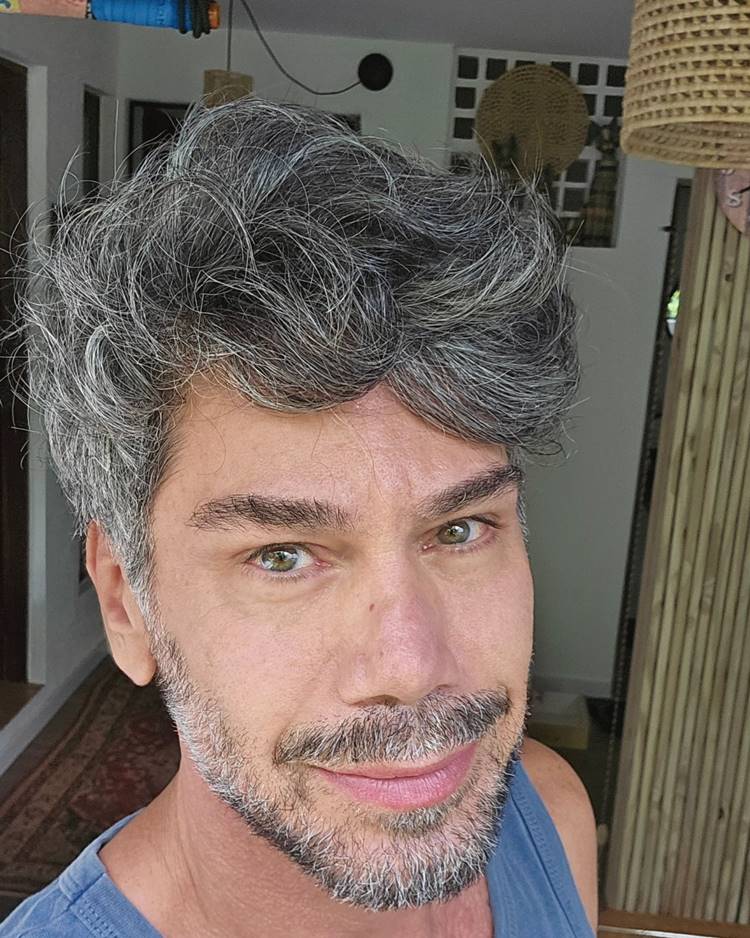 Homem com barba por fazer marcada, uma das tendências de 2024. Ele usa roupa azul e está em uma casa tirando selfie
