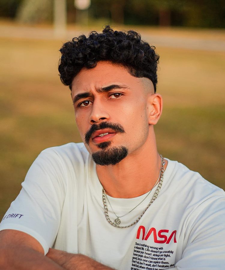Homem com barba estilo Van Dike, uma das tendências de 2024. Ele está com camiseta branca, correntes em fundo desfocado