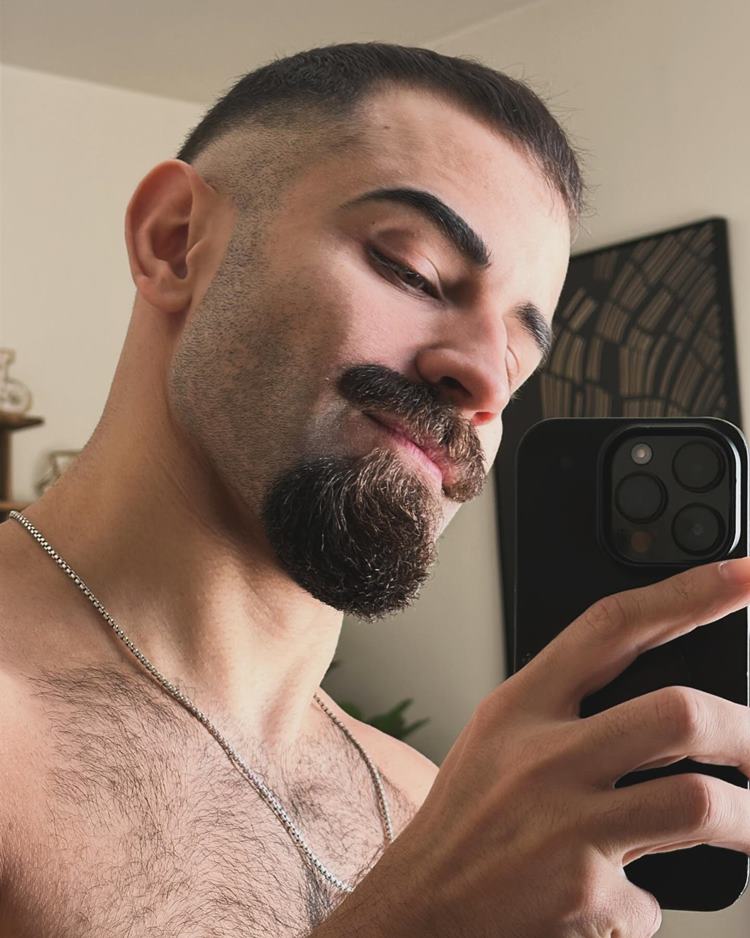 Homem com pelo do rosto estilo Van Dike, uma das tendências de 2024. Ele está sem camiseta, tirando selfie em espelho dentro de casa