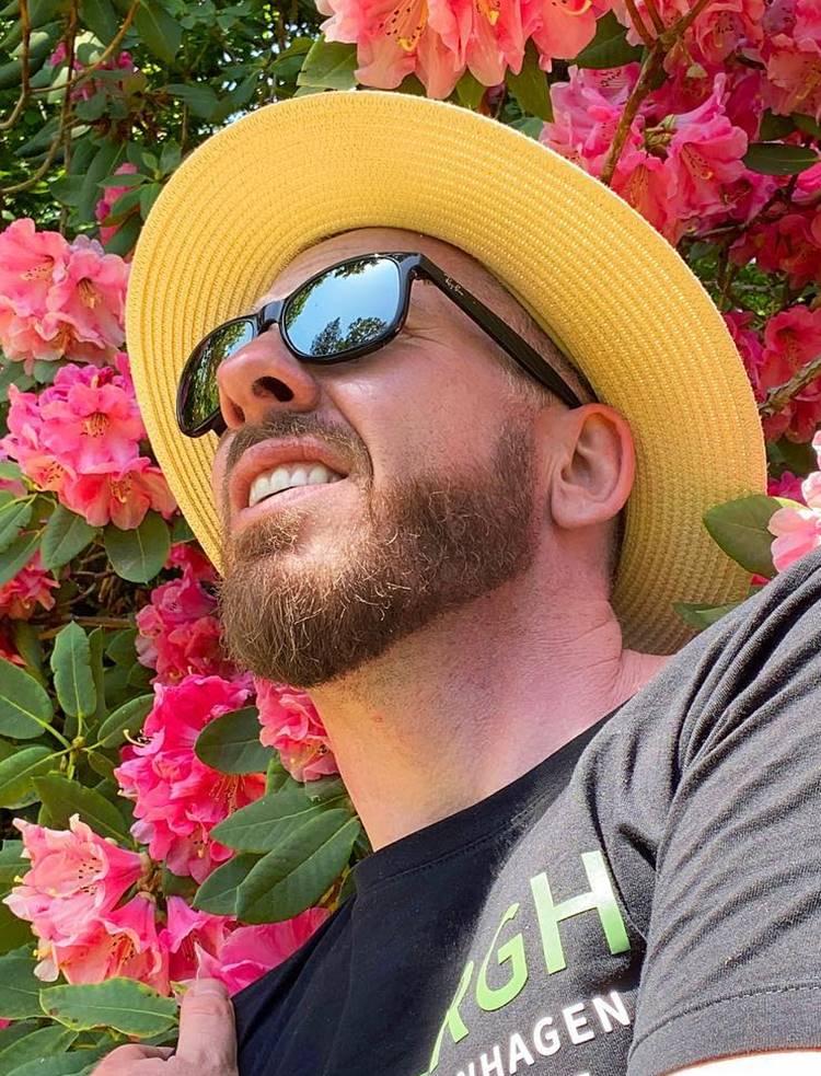 Homem com barba mais cheia no queixo, uma das tendências de 2024. Ele está com camisa preta, óculos de sol, chapéu e está em fundo de árvore com flores rosas