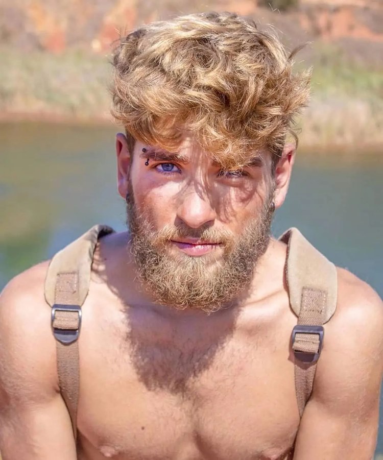Homem com barba mais cheia no queixo, uma das tendências de 2024. Ele está sem camisa, com mochila nas costas em fundo de águra com barranco