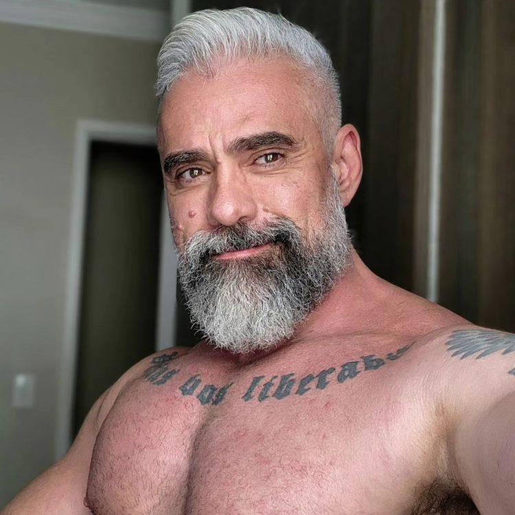 Homem com barba mais cheia no queixo, uma das tendências de 2024. Ele está sem camisa, tirando selfie em um cômodo e possui tatuagens no peito e braço
